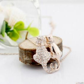 Rose gold necklace with intricate design on a wooden block, blurred glass container with greenery in the background.
